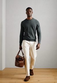 Brun lædertaske, mørkegrøn striktrøje, off-white bukser, brune ruskindsko. Neutral baggrund med lys træbelægning.