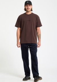 T-shirt en coton marron avec un col rond, arborant un petit logo noir. Associé à un pantalon en velours côtelé bleu marine et des chaussures à lacets noires.