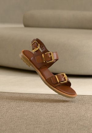 Brun lædersandal med to gyldne spænder og en gummisål, der svæver over en tekstureret beige tæppe.