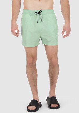 Maillots de bain vert clair avec une taille élastique et un cordon noir ; présente un petit logo sur le bas à l'avant. Porté avec des claquettes noires.