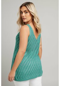 Tealfärgad stickad linne med V-formad ryggdesign, som har ett texturerat mönster. Bärs över vita byxor och visar en luftig silhuett.