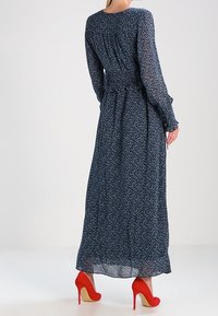 Maxi sukienka z ciemnoniebieskiego szyfonu w kwiatowy wzór, z długimi rękawami, talią przewiązaną paskiem i czerwonymi szpilkami; gładka faktura i zwiewny fason.