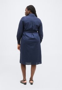 Γυναίκα με πλεξούδες στα μαλλιά, φοράει ένα μεσοφόρι πουκάμισο μίντι με μακριά μανίκια σε ναυτικό μπλε χρώμα και μαύρα παπούτσια με χαμηλό τακούνι, στέκεται μπροστά σε λευκό φόντο.