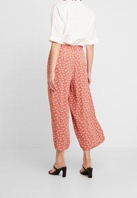 Pantalons à jambes larges de couleur corail avec un motif floral blanc. Le tissu semble léger avec une coupe décontractée. Associés à des sandales à talons blocs noires.