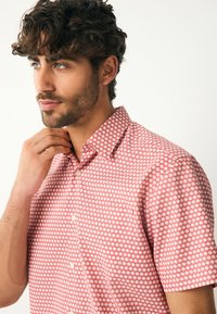 Chemise rouge à manches courtes avec un motif floral blanc, col boutonné et tissu en coton lisse. Présente une coupe classique et une couture soignée.
