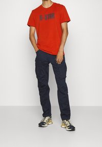 Röd bomullströja med "G-STAR"-logotyp, matchad med marinblå cargobyxor med sidofickor och en avslappnad passform. Svarta och gula träningsskor.