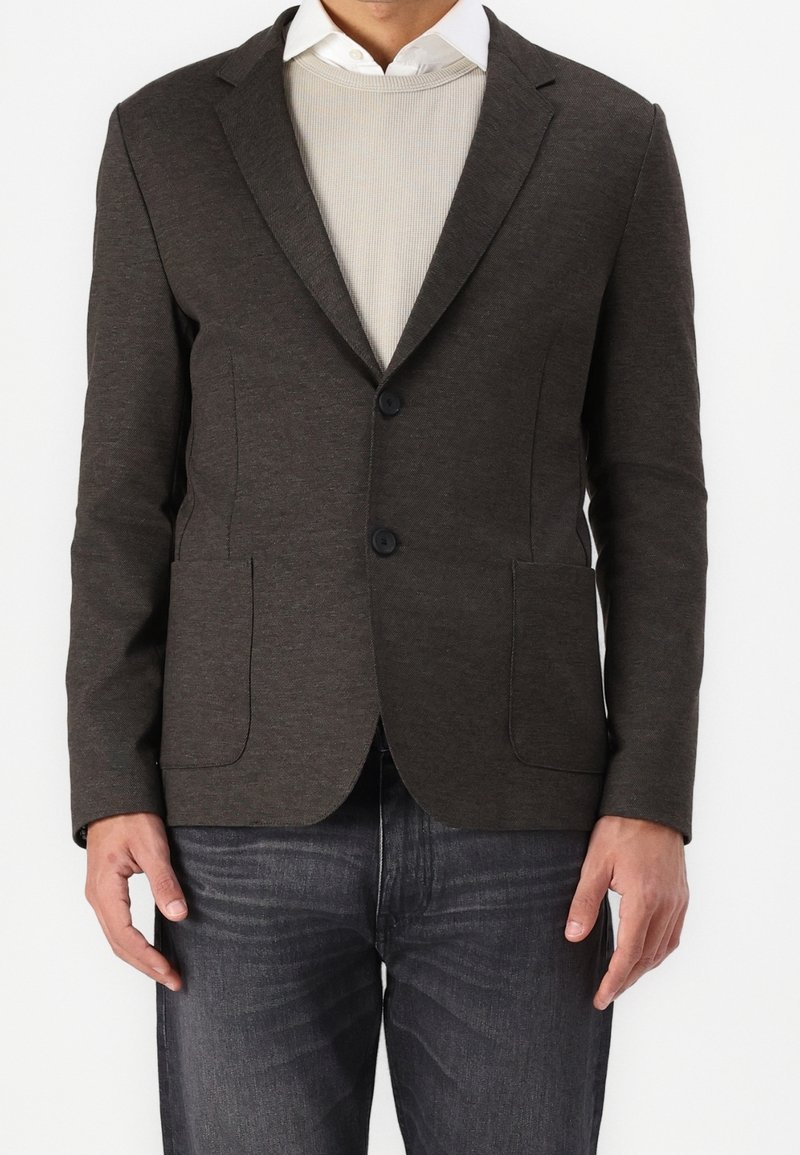 Donkergrijze blazer van gladde stof, met twee voorzakken, een enkelvoudige knoopsluiting en twee knopen. Gedragen over een lichte trui.