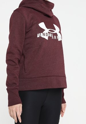 Person iført en burgunder Under Armour hættetrøje med høj krave og sorte leggings, stående mod en ensfarvet hvid baggrund.