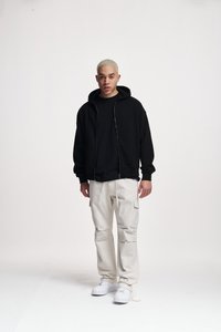 Felpa nera con zip sopra una maglietta nera, abbinata a pantaloni cargo color crema e sneakers bianche. Sullo sfondo c'è un bianco semplice.