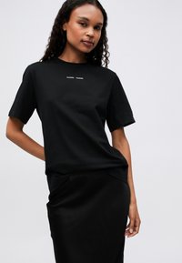 Black cotton t-shirt with short sleeves, featuring a small white logo on the chest, styled with a matching black satin skirt.