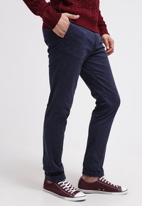 Person trägt marineblaue Hose, weinrote Sneakers mit weißen Schnürsenkeln und einen weinroten Zopfpullover, eine Hand in der Tasche.