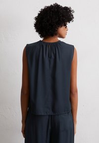 Frau mit lockigem Haar, die eine ärmellose dunkelblaue Bluse und passende Hosen trägt, von hinten vor einer weißen strukturierten Wand gezeigt.