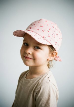 Junges Kind mit heller Haut und grünen Augen, trägt ein beiges Hemd und eine rosa Mütze mit Kirschmuster, lächelt sanft vor hellem Hintergrund.