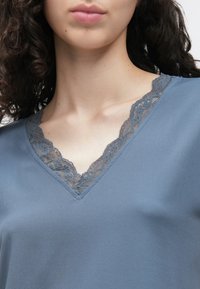 Sininen V-kaula-aukollinen paita, valmistettu sileästä kankaasta, jossa koristuksena harmaa pitsi kaula-aukon ympärillä, lyhyet ja laskeutuvat hihat.