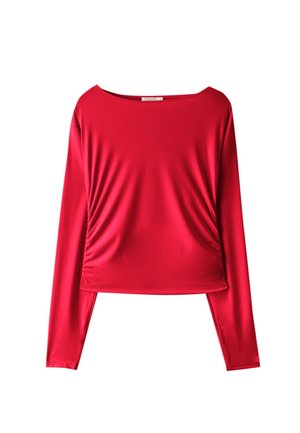 Femme portant un crop top rouge à manches longues et un pantalon, tenant une tasse noire et une veste blanche, debout à côté de poutres métalliques rouges sur fond gris.