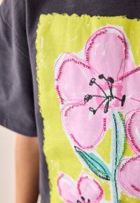 Pilkas marškinėlis su dideliu gėlių dizainu: rausvos gėlės, žali lapai ir ryški gelsva kraštinė, papuošta karoliukais ir tekstūromis.