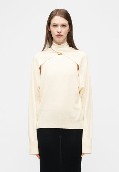 Femme portant un pull crème avec un col superposé unique et une jupe noire, debout devant un fond blanc uni.