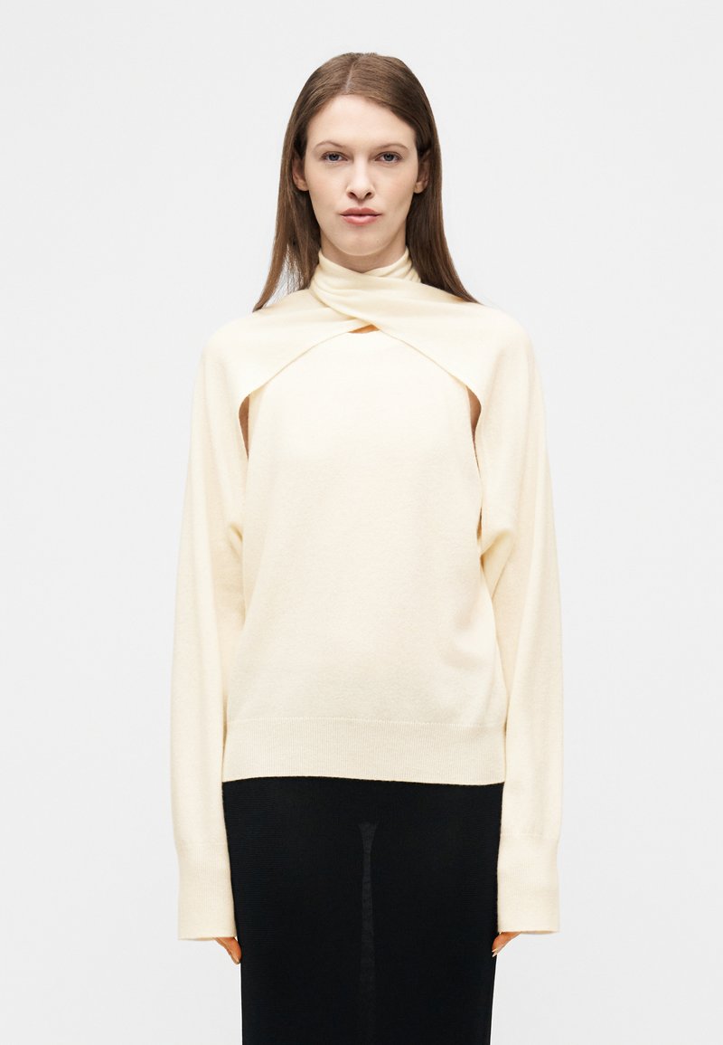 Γυναίκα με γκρι κρεμ πουλόβερ με μοναδικό σχεδιασμό γιακά που αλληλοεπικαλύπτεται και μαύρη φούστα, στέκεται μπροστά σε απλό λευκό φόντο.