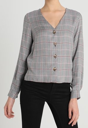 Femme portant un chemisier à carreaux gris à manches longues avec col en V, associé à un pantalon noir ajusté.