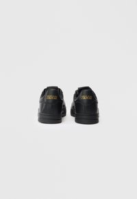 Par črnih nizkih športnih copat z zlatim logotipom "Versace Jeans Couture" na peti, prikazanih s hrbtne strane na enobarvni beli podlagi.