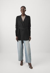Svart, bältetrenchcoat i mjukt tyg, med en notchad krage och långa ärmar. Bärs över ljusblå, lösa jeans och högklackade skor.