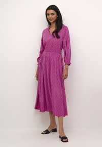 Duga ružičasta haljina s v-izrezom, nabranim strukom i elastičnim rukavima, s geometrijskim uzorkom i lepršavom suknjom. Uparen s crnim cipelama.