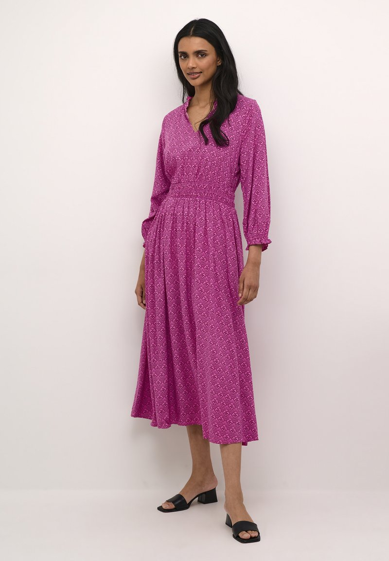 Duga ružičasta haljina s v-izrezom, nabranim strukom i elastičnim rukavima, s geometrijskim uzorkom i lepršavom suknjom. Uparen s crnim cipelama.