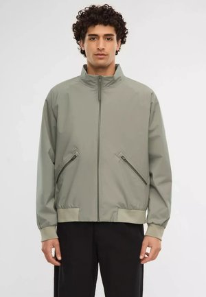 Man met krullend haar, gekleed in een lichtgrijs jack met ritszakken en een zwarte broek, staand tegen een effen witte achtergrond.