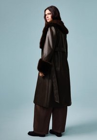 Dlouhý tmavě hnědý kožený kabát s koženým límcem a manžetami, s opaskem v pase, a volně střižené hnědé kalhoty, umístěný na modrém pozadí.