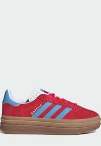 Röd mockasneaker med blå accenter, tre vita ränder, texturerad vit tunga och en kraftig beige sula. Har "Gazelle"-märkning.