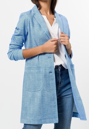 Femme ajustant un blazer texturé bleu clair sur un chemisier blanc à col en V et un jean bleu, sur un fond blanc uni.