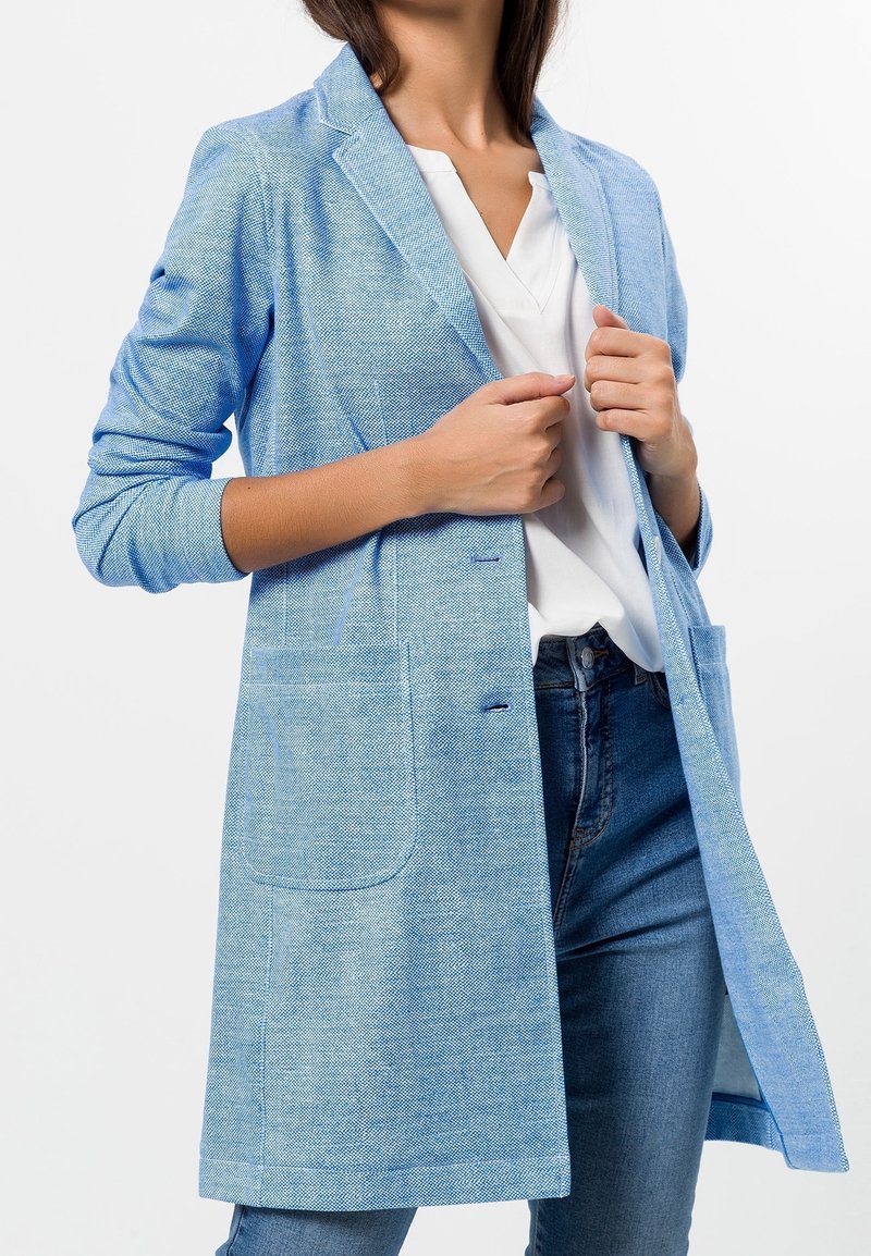 Femme ajustant un blazer texturé bleu clair sur un chemisier blanc à col en V et un jean bleu, sur un fond blanc uni.
