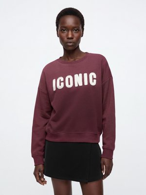 Schwarze Frau trägt ein bordeauxfarbenes Sweatshirt mit dem Wort "ICONIC" in weißen Buchstaben und einen schwarzen Minirock vor weißem Hintergrund.