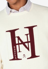 Suéter de punto color crema con un gran diseño en marrón "H" y "N" con el texto "HARPER" debajo, acompañado de un gráfico de un sombrero cruzado y herramientas.