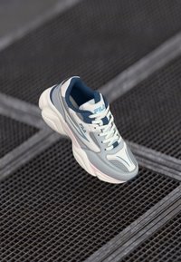 Zapatilla gris y blanca con lengüeta acolchada y cierre con cordones. Presenta paneles lisos y texturizados, acentos en azul y una suela rosa.