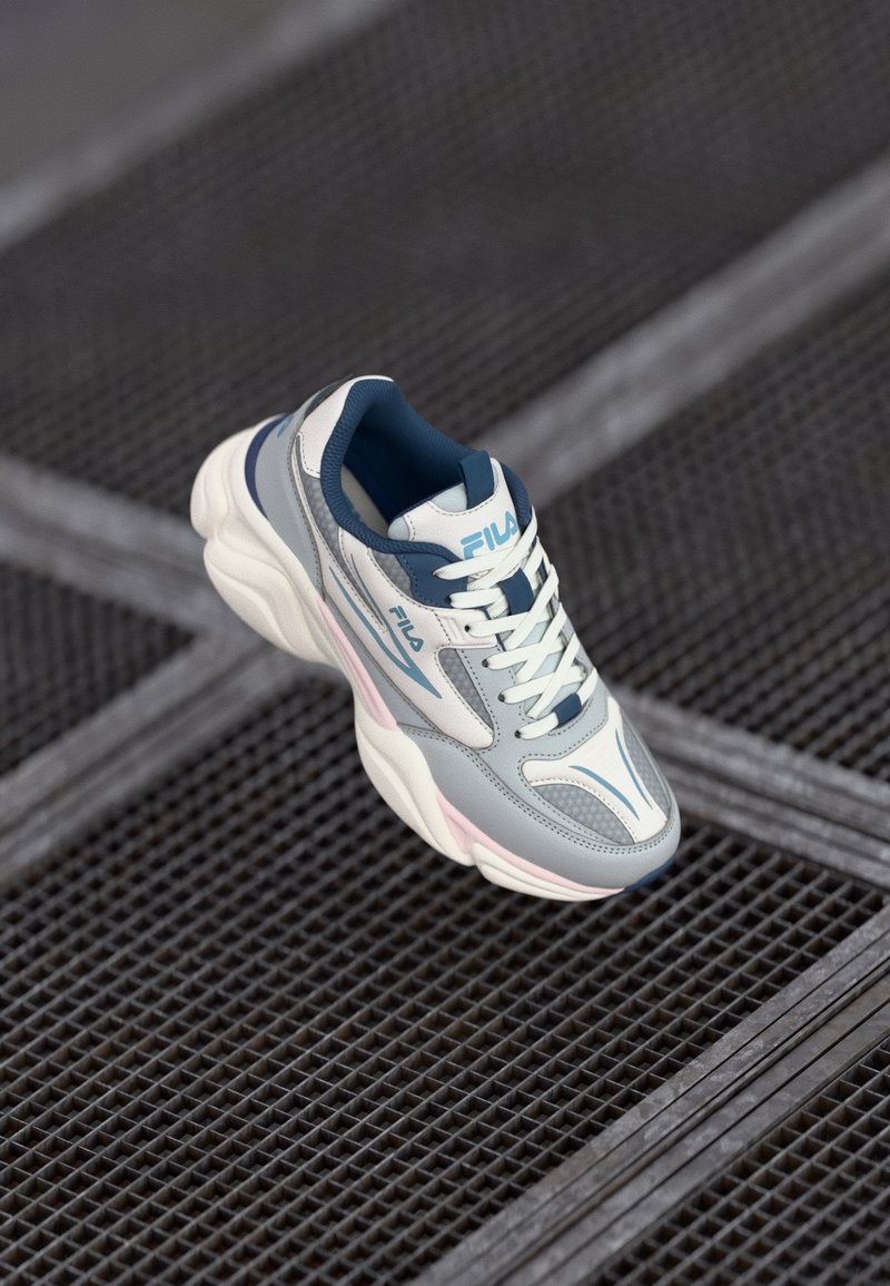 Zapatilla gris y blanca con lengüeta acolchada y cierre con cordones. Presenta paneles lisos y texturizados, acentos en azul y una suela rosa.