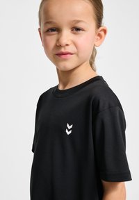 T-shirt noir à manches courtes fabriqué en matériau doux, avec un logo en chevron blanc sur la partie gauche de la poitrine. Texture lisse.