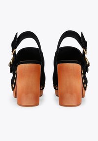 Czarne zamszowe sandały na obcasie z drewnianym blokiem, regulowanym paskiem wokół kostki i złotymi akcentami. Dekoracyjne ćwieki wzdłuż boku.