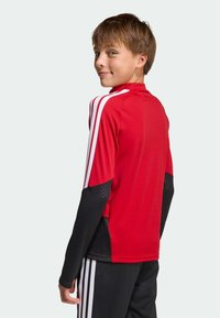 Κόκκινη αθλητική φανέλα με μαύρα μακριά μανίκια, με λευκές κάθετες ρίγες στους ώμους και υφασμένο μαύρο πάνελ.