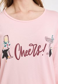 Rosa kortärmad t-shirt med "Cheers!" i vinröd färg, med illustrationer av glas och pingviner på varje sida. Slät bomullstextur.