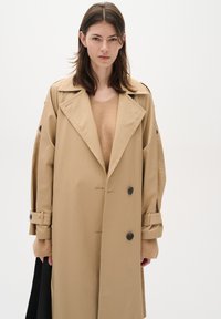 Beige trench coat with a double-breasted design, black buttons, wide lapels, and pronounced sleeves. Worn over a light brown sweater.
