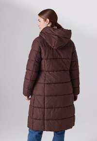 Pruun polsterdatud pikk jope kapuutsiga, vertikaalse tikandiga, pikkade varrukatega ja lõtva istuvusega, koos siniste teksadega.