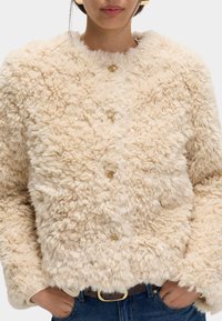 Kunstpelzjacke in einem sanften Beigeton, mit rundem Ausschnitt, goldenen Knöpfen und einer flauschigen Textur für Wärme und Komfort.