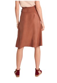 Donna che indossa una gonna marrone ruggine al ginocchio con una cintura larga, abbinata a scarpe espadrillas bordeaux, vista da dietro.