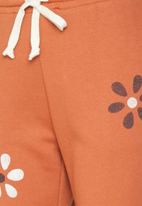 Calças de fato de treino cor ferrugem com um cordão branco, apresentando padrões florais em preto e branco impressos na parte inferior. Tecido suave e texturizado.