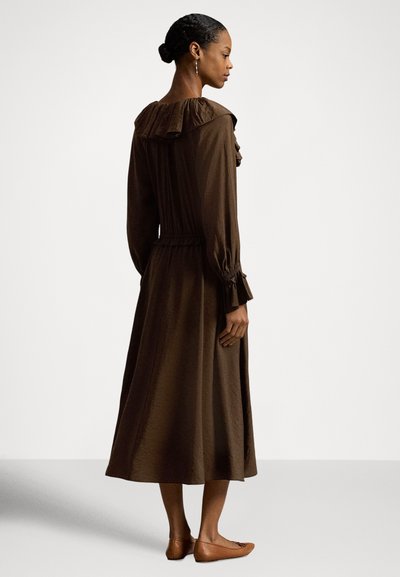 Ruda, ilgomis rankovėmis suknelė su raukiniais kaklo linijoje, prigludusia liemeniu ir minkšta tekstūra, derinama su rudais butais, kuriuose vyrauja minimalistinis dizainas.
