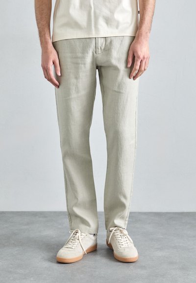 Pantalons en lin gris clair avec une coupe droite, présentant un design simple. Associés à des baskets à lacets blanches cassées et un haut de couleur neutre.