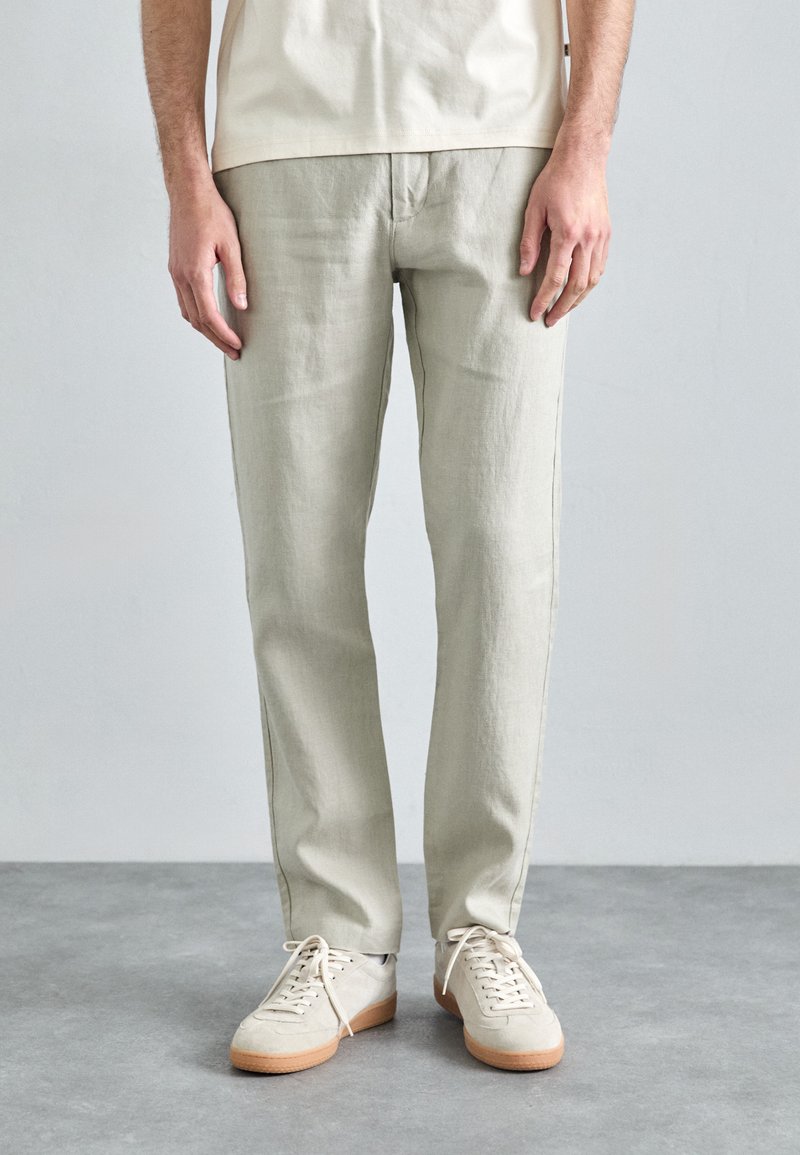 Pantalons en lin gris clair avec une coupe droite, présentant un design simple. Associés à des baskets à lacets blanches cassées et un haut de couleur neutre.