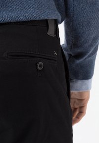 Černé chinos kalhoty s zadní kapsou na knoflík, hladká bavlněná látka a úzký střih. Viditelné prošití dodává detail.