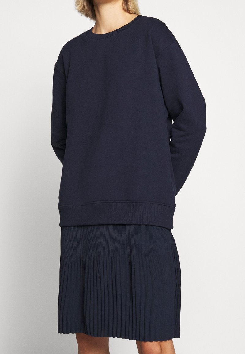 Personne portant un sweat-shirt bleu marine à manches longues et une jupe plissée bleu marine sur un fond uni clair.